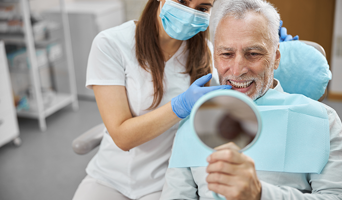 Older man sitting in a dental chair looking in a handheld mirror at his new dental implants from Matsui Dental in New York, NY